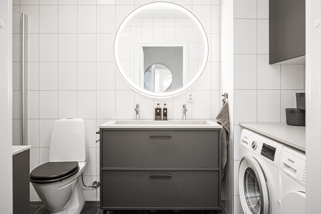 Badrum med grå kommod, rund spegel och tvättmaskin