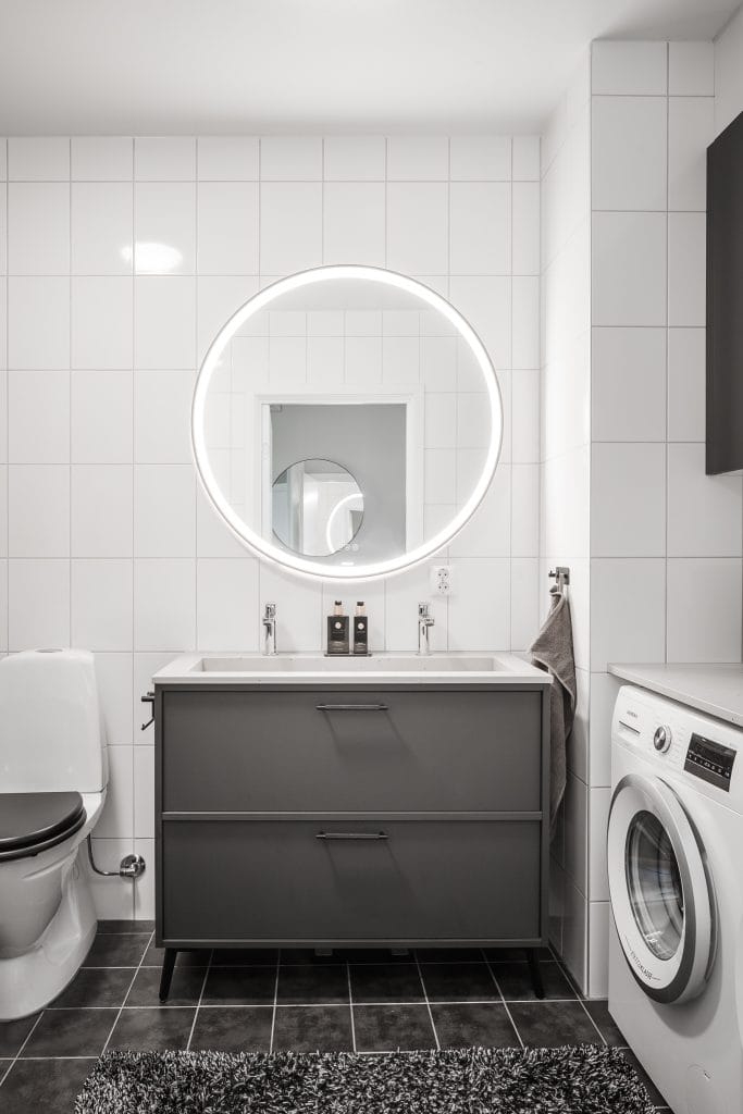 Badrum med grå kommod, rund spegel och tvättmaskin